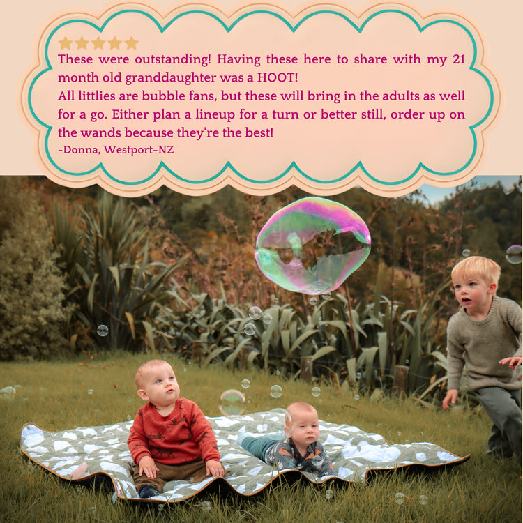 2 little children sitting on a picnic blanket in a meadow surrounded by bubbles and 1 little boy chasing a giant bubble.