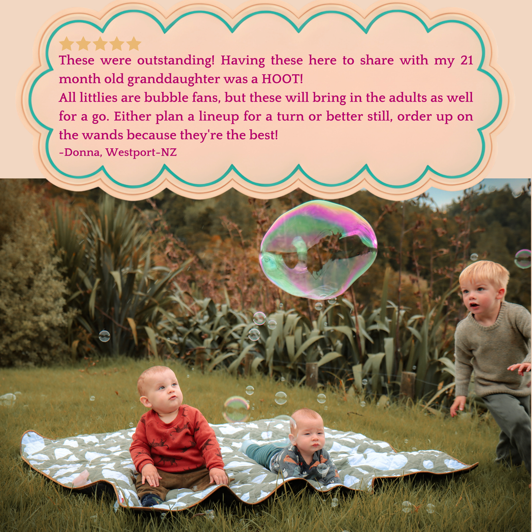 2 little children sitting on a picnic blanket in a meadow surrounded by bubbles and 1 little boy chasing a giant bubble.